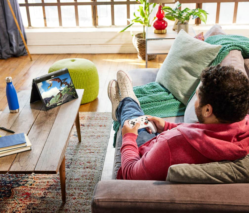 A person sits on a sofa with their feet up and plays video games on a Chromebook using a game controller.